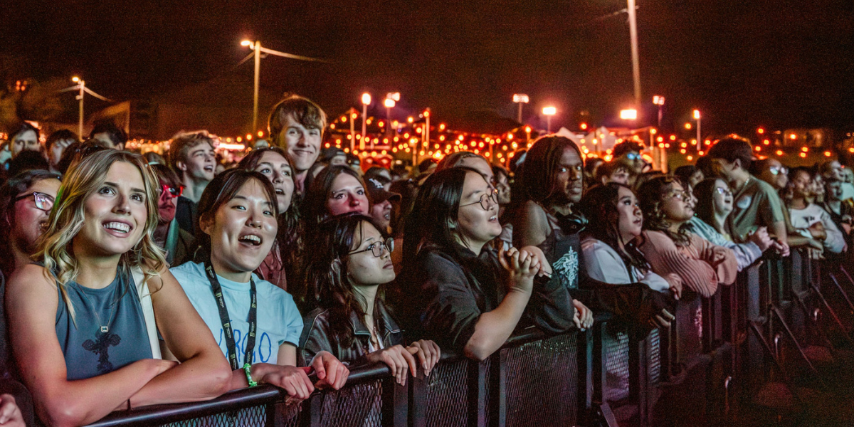 A crowd of people excitedly watching an outdoor nighttime event behind barriers, with decorative lights in the background.