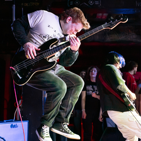 Musicians performing on stage, one jumping with a bass guitar.